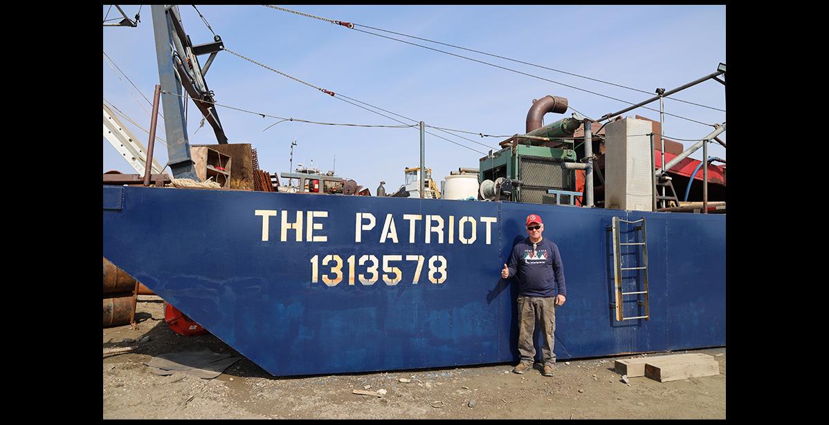 Photo by Colin Warren TRUE PATRIOT— Retired Marine Veteran Henry Brosnaham stands in front of his large dredge as he is preparing for the gold mining season in the dry docks, at the Port of Nome.
